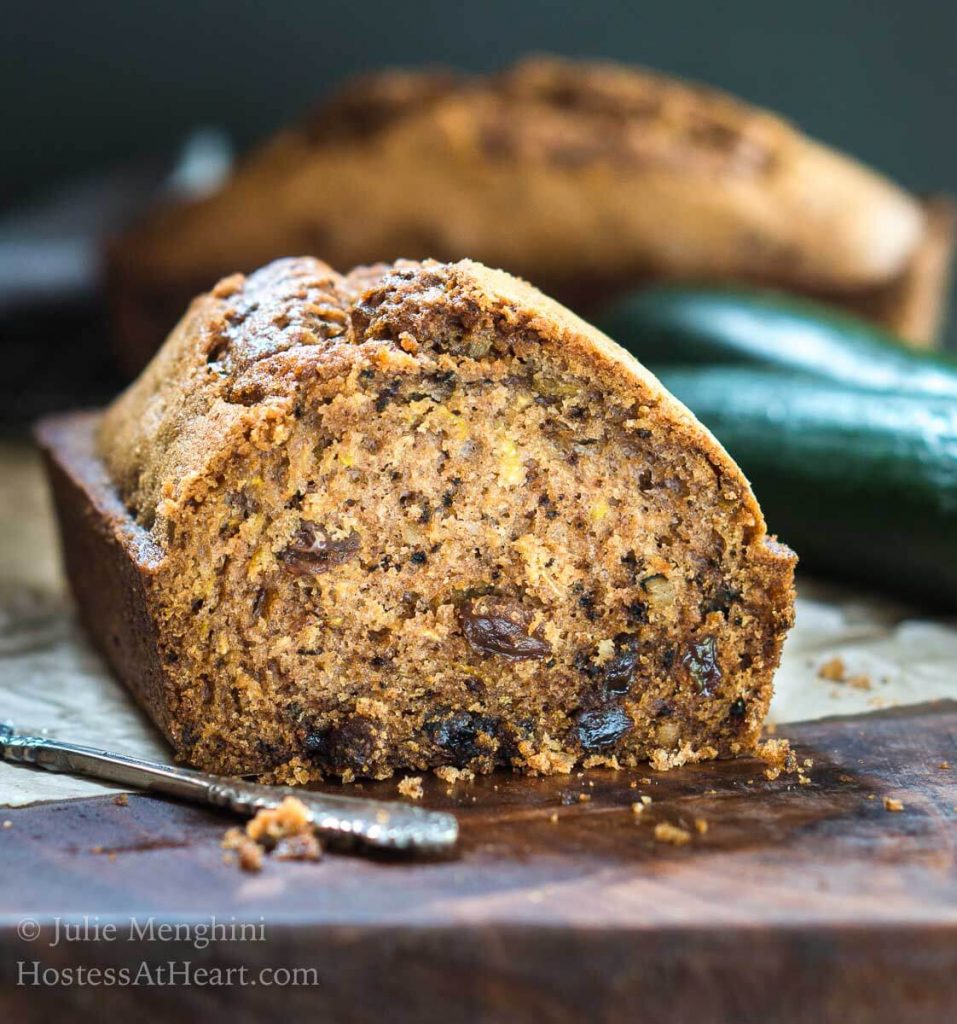 Front view of a sliced loaf of Zucchini bread sitting on a wooden cutting board. A full loaf and raw zucchini sit in the background.