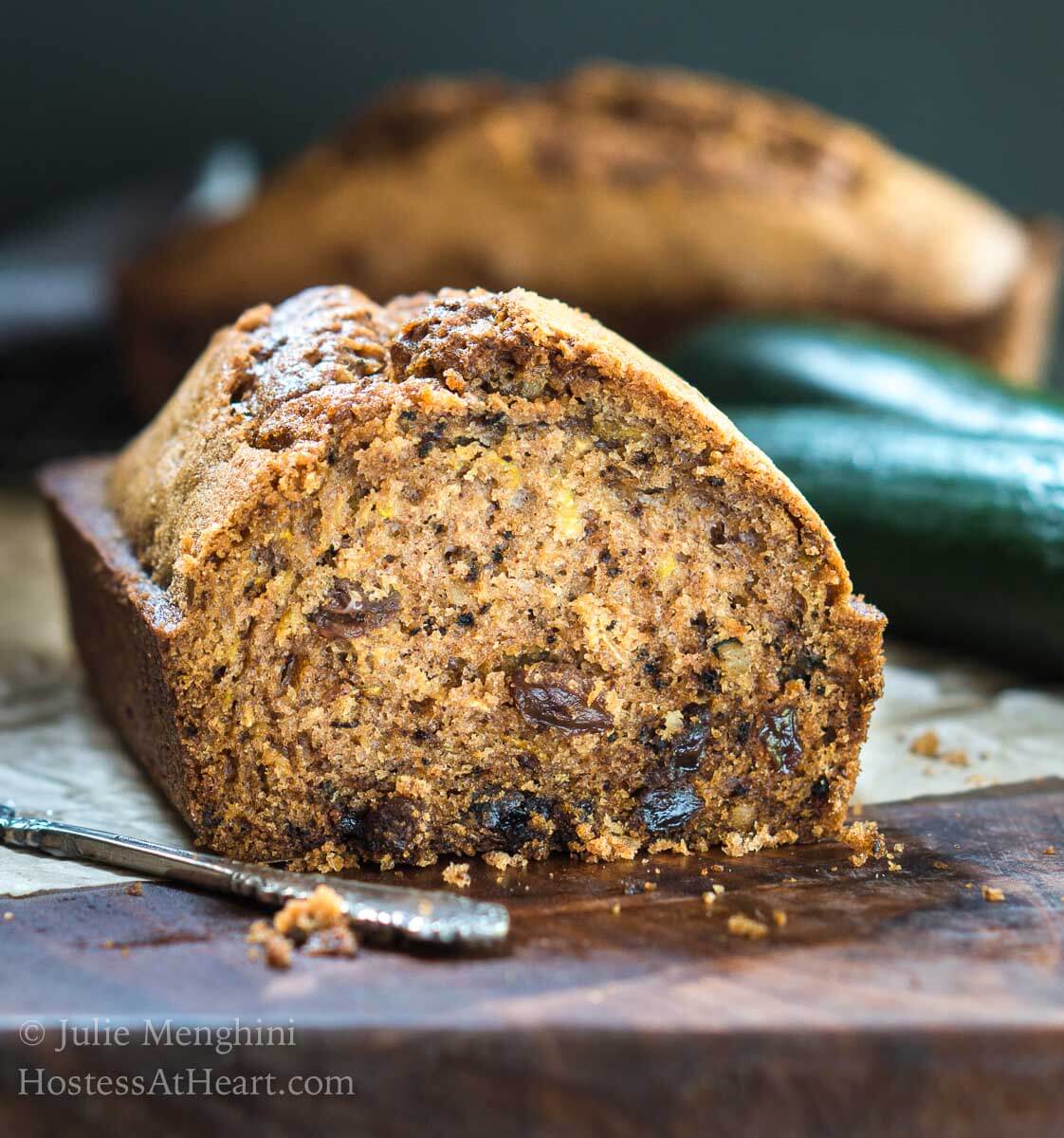 Front view of a sliced loaf of Zucchini bread sitting on a wooden cutting board. A full loaf and raw zucchini sit in the background.