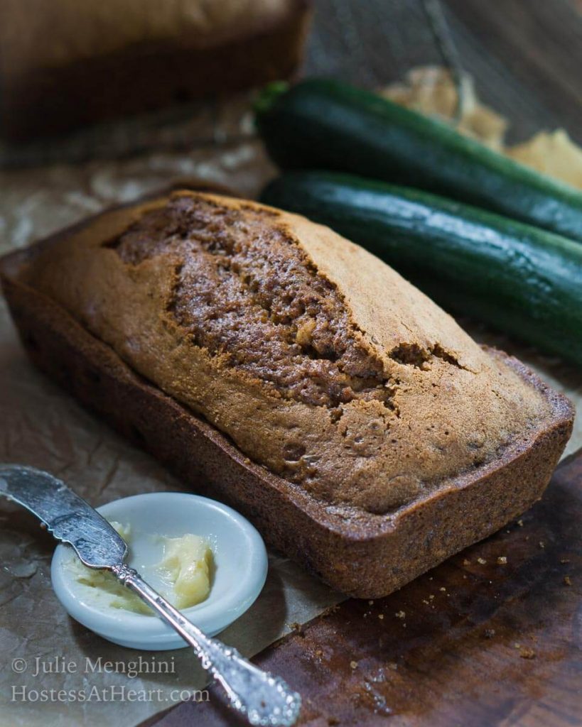 Top angle view of a loaf of zucchini bread. A small white dish holding butter and a knife and two raw zucchini sit to the side.