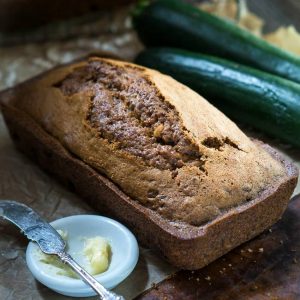 Top angle view of a loaf of zucchini bread with a white dish holding butter and a knife next to it. Raw zucchini sit next to the bread.