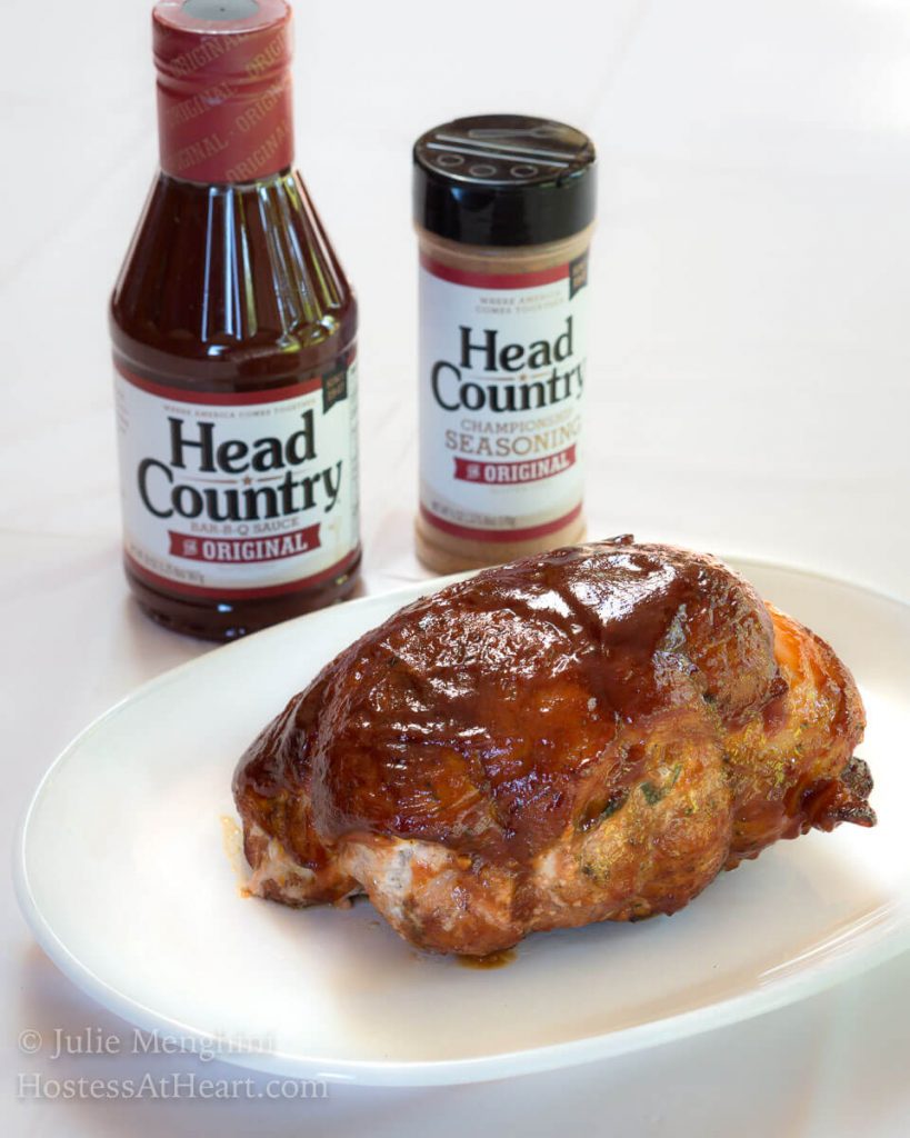 A grilled pork tenderloin glazed in bbq sauce sitting on a white platter. Head Country BBQ and seasoning sit in the background.