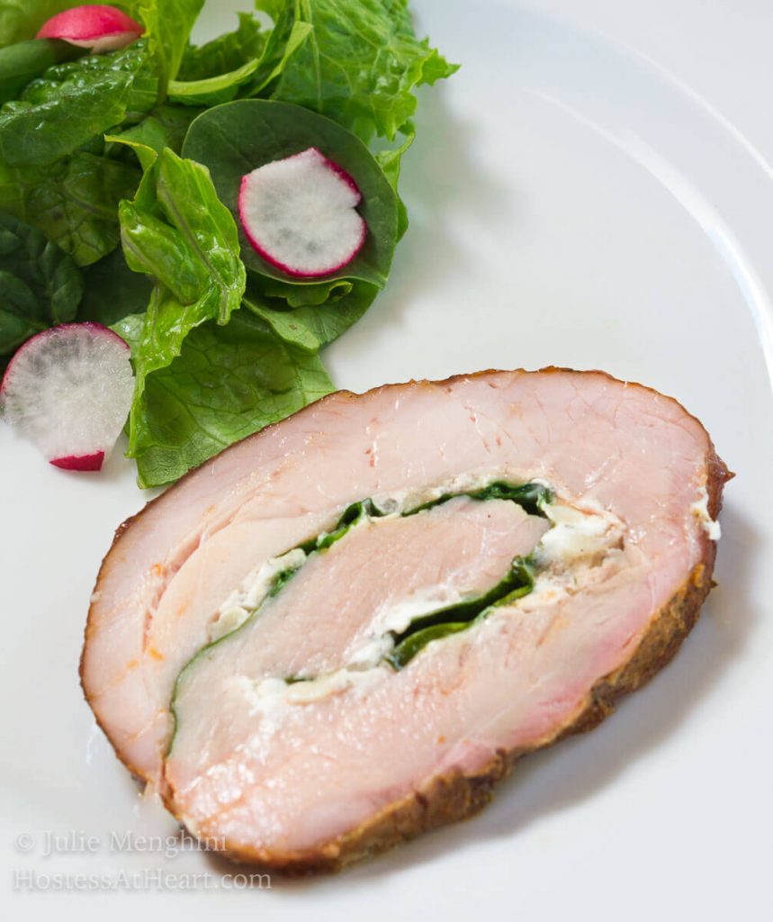 Top-angle view of a slice of pork tenderloin that\'s been stuffed with goat cheese and spinach, tied and grilled over a white plate next to a lettuce salad.