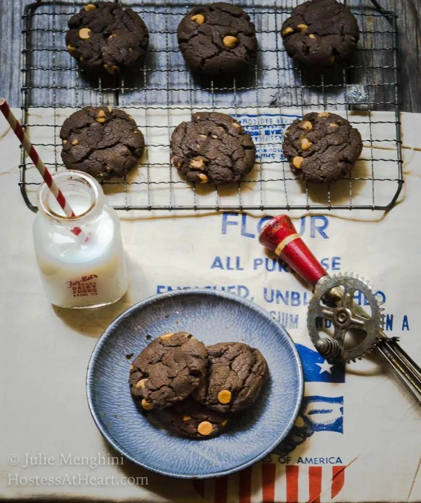 Top-down photo of Chocolate cookies dotted with peanut butter chips sit on a cooling rack over a flour sack. A blue plate holding two cookies is in the front next to a jar of milk and an antique hand mixer sit to the side.