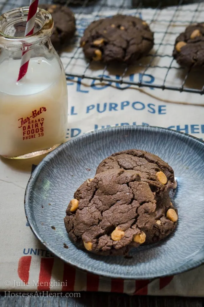 Two Chocolate cookies dotted with peanut butter chips sit in a blue plate over a flour sack. A jar of milk and a cooling rack full of cookies sit in the background.