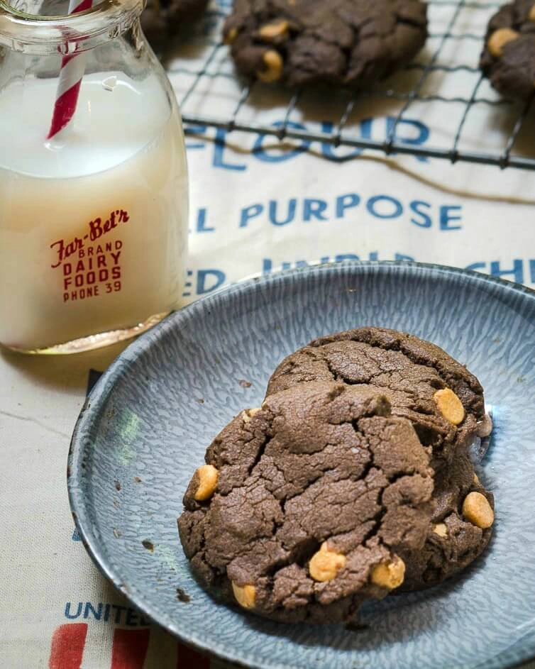 Three Chocolate Peanut Butter Chip cookies sitting on a blue enamelware plate. A jar of milk sits next to the plate and a cooling rack filled with cookies sits in the background. 