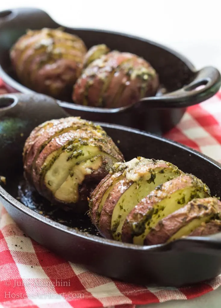 Two cast-iron casserole dishes holding red potatoes that have been sliced and loaded with garlic basil sauce over a red checked tablecloth.