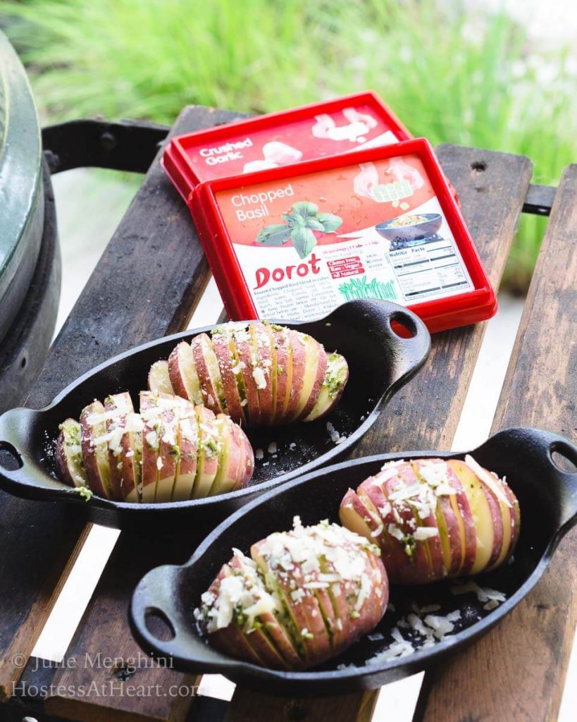 Two cast-iron casserole dishes holding uncooked red potatoes that have been sliced and loaded with garlic basil sauce over a red checked tablecloth. Packages of Dorot Garlic sit in the background.