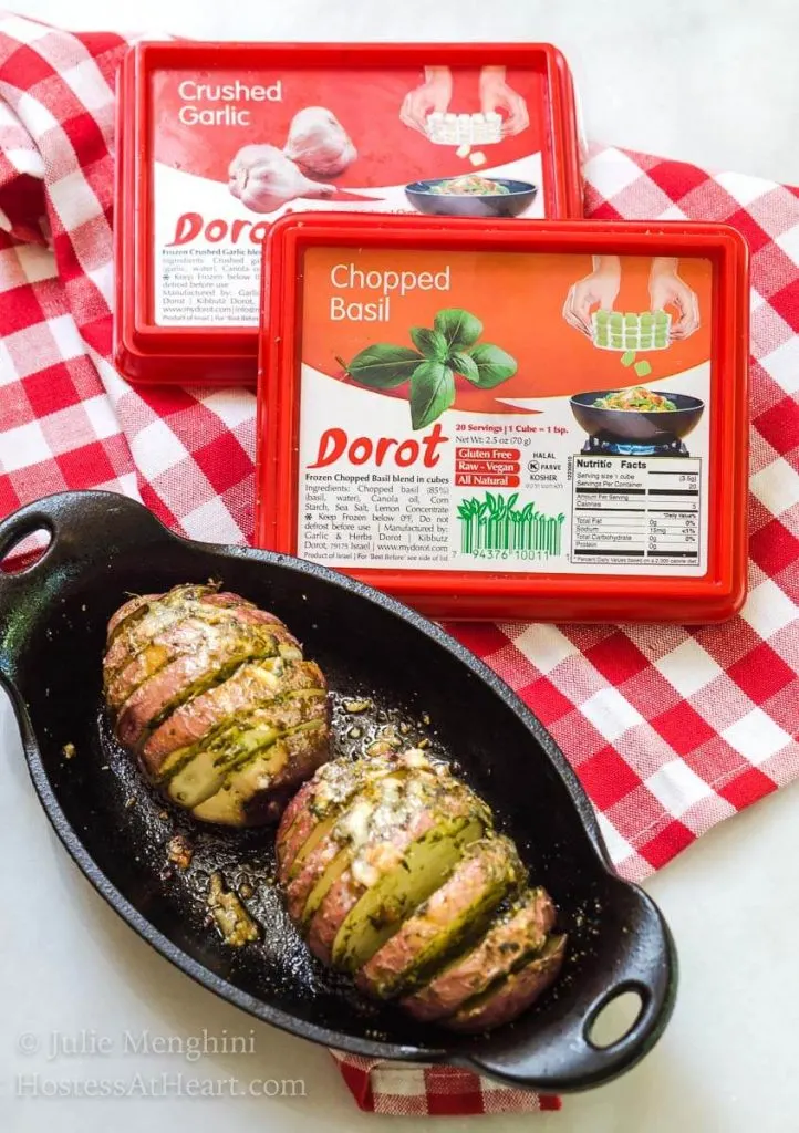 A cast-iron casserole dish holding a Hasselback red potato seasoned with basil garlic basil sauce. The Dorot Basil and Garlic packages sit in the back over a red checkered napkin.
