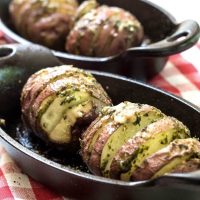 Two cast-iron casserole dishes holding red potatoes that have been sliced and loaded with garlic basil sauce over a red checked tablecloth.