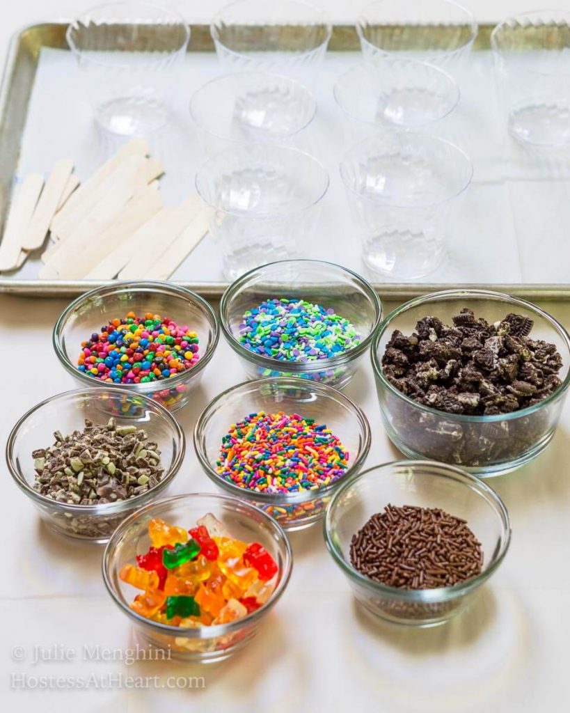 Dishes of multiple colored sprinkles for making Ice Cream Pops. Empty plastic cups sit in the background.