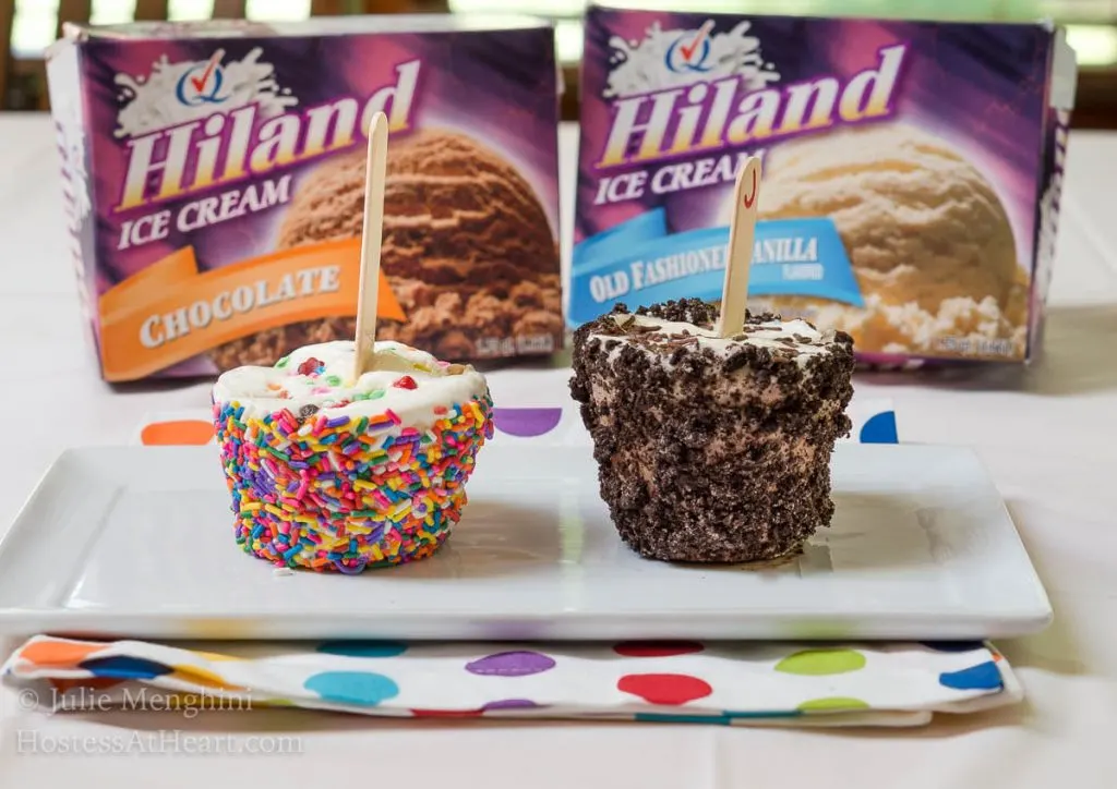 Two ice cream pops rolled in rainbow sprinkles and cookie crumbs with popsicle sticks in the center sitting on a white plate. Highland Ice Cream containers sit in the background.