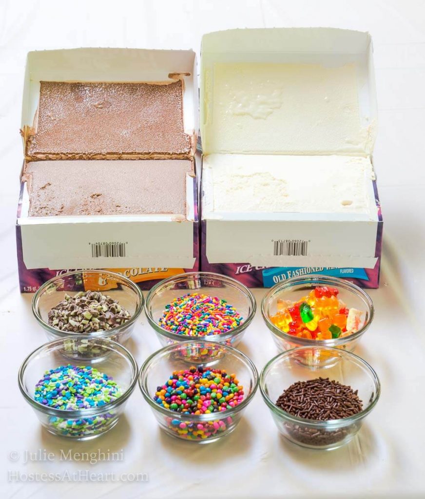 Two open containers of vanilla and chocolate ice cream and dishes of multiple colored sprinkles for making Ice Cream Pops sit on a white table.