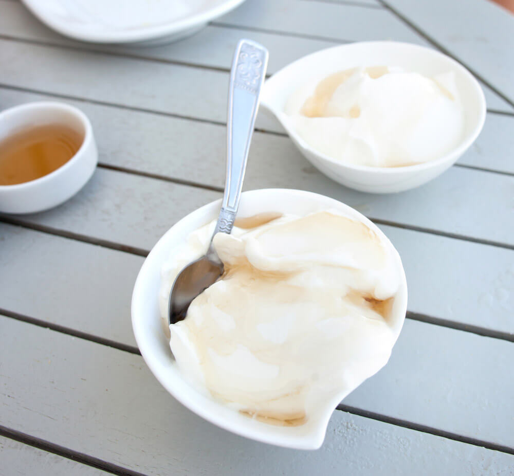 A white dish of yogurt drizzled with honey. White bowls of yogurt and honey sit in the background.
