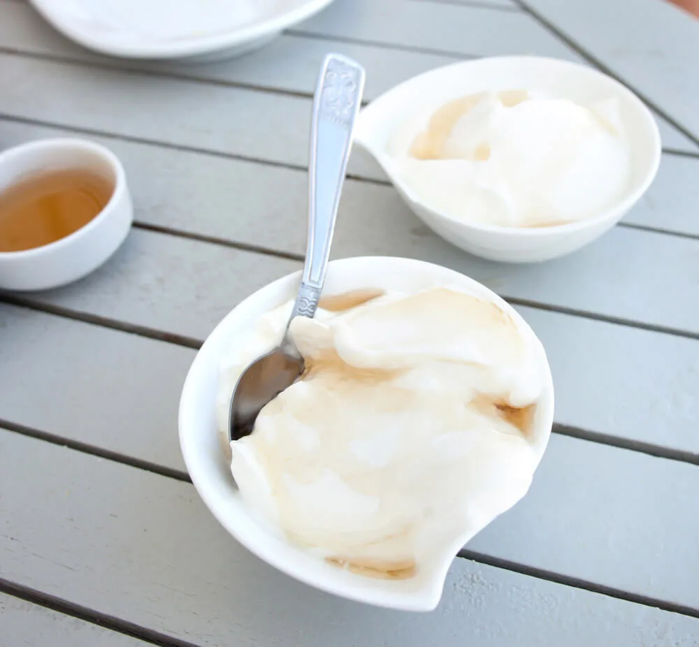 A white dish of yogurt drizzled with honey. White bowls of yogurt and honey sit in the background.