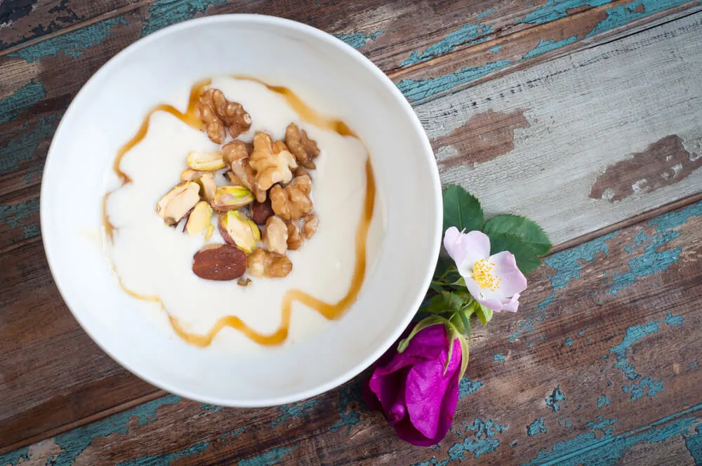 A shite bowl filled with yogurt and drizzled with honey and nuts sits over a painted wooden table. Two pink blossoms sit next to the bowl. 