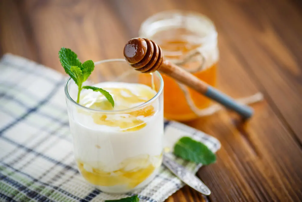 A glass cup holding yogurt blended with honey and garnished with mint over a wooden background. A jar of honey sits in the background.