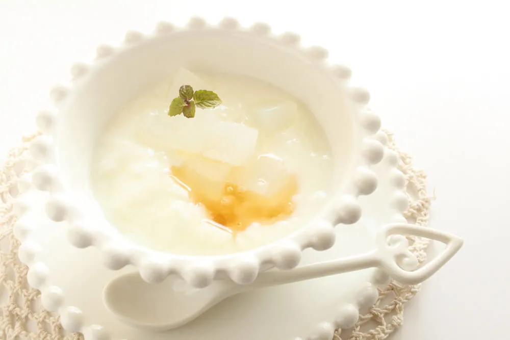 A white dish filled with yogurt that\'s been drizzled with honey. A matching spoon sits on the plate holding the dish.