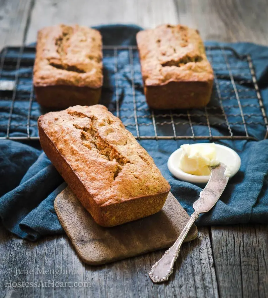 A loaf of banana bread sitting on a wooden butter paddle with a white dish of butter and knife beside it. Two loaves sit on a cooling rack over a blue napkin in the background.