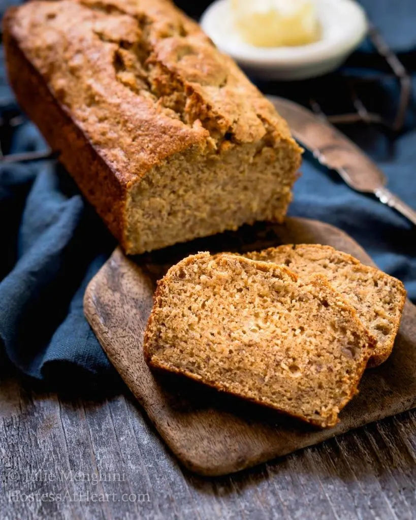 A side view of a loaf of banana bread sitting on a wooden butter paddle with slices laying in the front. A blue napkin sits in the back.