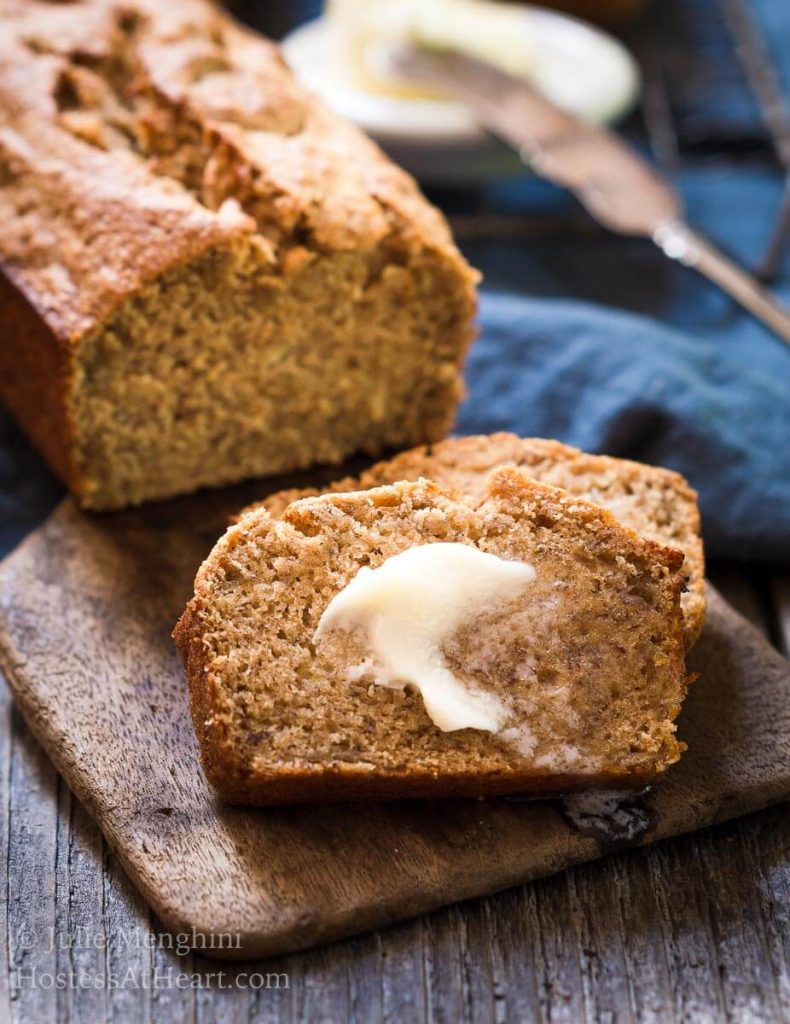 A side view of a loaf of banana bread sitting on a wooden butter paddle with slices laying in the front covered with melted butter. A blue napkin sits in the back.