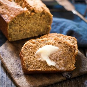 A side view of a loaf of banana bread sitting on a wooden butter paddle with slices laying in the front. A blue napkin sits in the back.