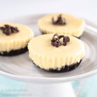 Mini Cheesecakes have an Oreo crust, white cheesecake filling and topped with chocolate shavings. They sit on a small cake plate over a blue napkin. 