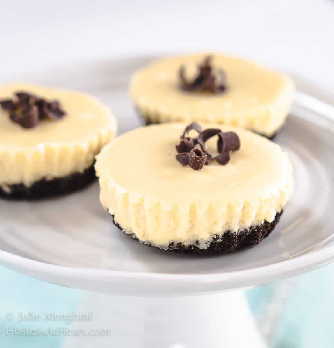 Mini Cheesecakes have an Oreo crust, white cheesecake filling and topped with chocolate shavings. They sit on a small cake plate over a blue napkin. 