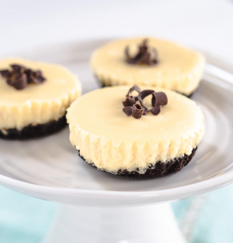 Side view of 3 Mini Cheesecakes on a white pedestal plate. They are layered with a chocolate oreo crust, a white chocolate filling, and chocolate shavings over the top.