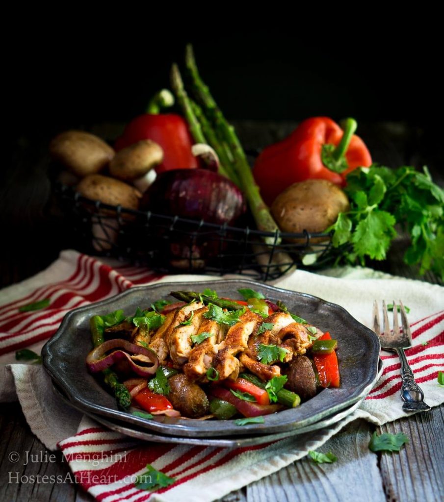 Fajita vegetables topped with slices of chicken on a red striped napkin. Raw peppers, asparagus, mushrooms, and cilantro sit in the background.