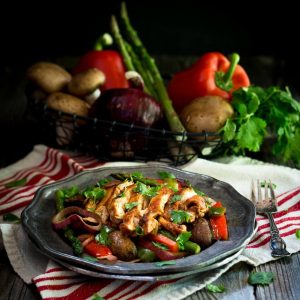 A metal plate filled with sliced chicken red peppers, mushrooms, and asparagus mixed with spices sitting on a red striped towel. Fresh vegetables sit in a metal basket in the background.