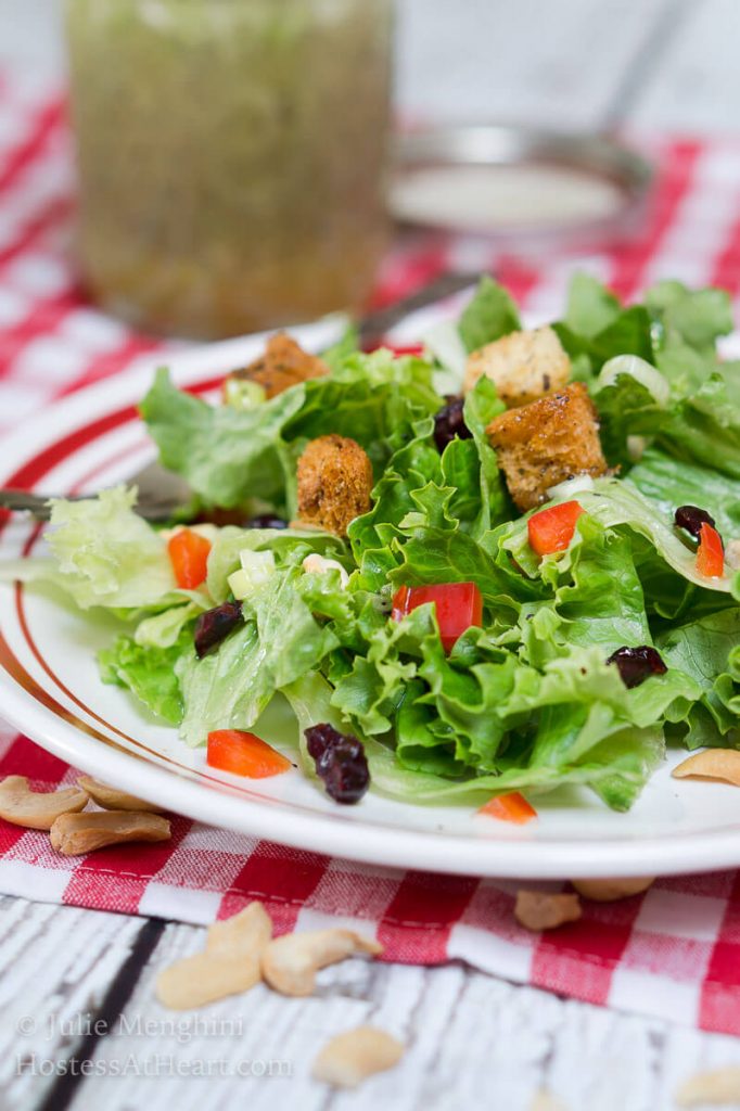A white plate with a red rim filled with a salad that\'s dotted with green onion, craisins, croutons, and nuts. A jar of vinaigrette over a red-checked tablecloth sits in the background.