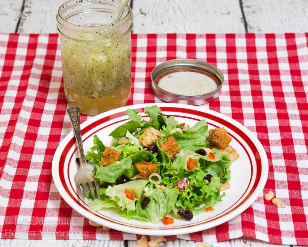 A white plate with a red rim filled with a salad that\'s dotted with green onion, craisins, croutons, and nuts. A jar of vinaigrette over a red-checked tablecloth sits in the background.