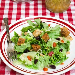 A white plate with a red rim filled with a salad that's dotted with green onion, craisins, croutons, and nuts. A jar of vinaigrette over a red-checked tablecloth sits in the background.