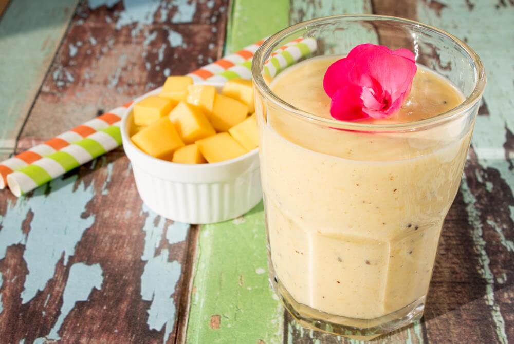 A glass of Mango Smoothie with a white dish of diced mango sitting behind it on top of a painted wooden tabletop.