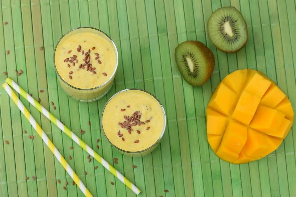 A mango and kiwi cut in half next to two smoothies on a green wood board