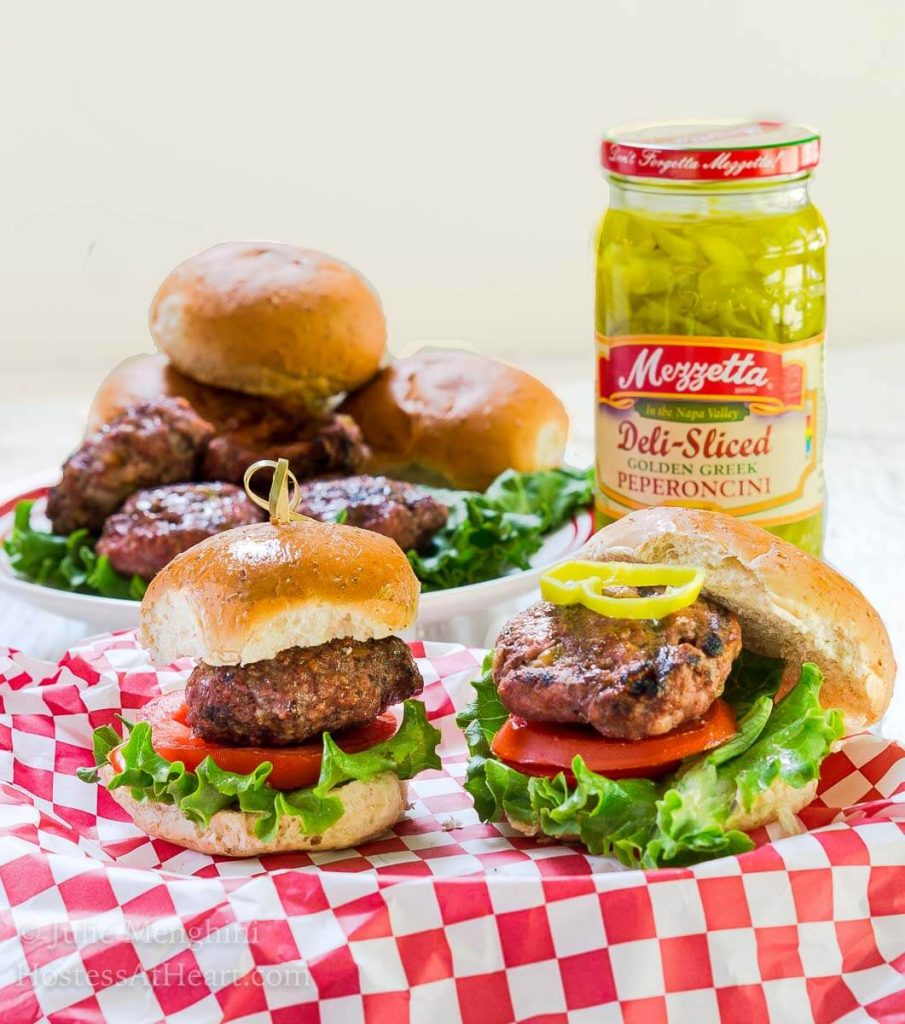 Side view of a hamburgers that\'s been stuffed with Pepperjack cheese and pepperoncini sitting over a slice of tomato and lettuce between a bun. A skewer secures the bun to the burger. over a red-checked cloth. A jar of pepperoncini sits in the back next to a platter of burgers.