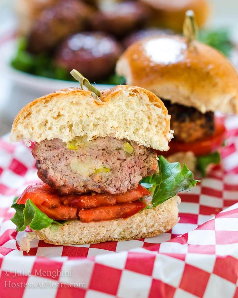 Side view of a hamburger cut in half that\'s been stuffed with Pepperjack cheese and pepperoncini sitting over a slice of tomato and lettuce between a bun. A skewer secures the bun to the burger. over a red-checked cloth.