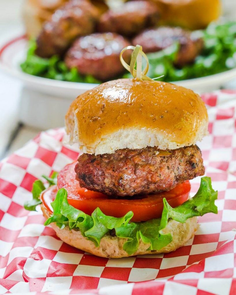 Side view of a hamburger that\'s been stuffed with Pepperjack cheese and pepperoncini sitting over a slice of tomato and lettuce between a bun. A skewer secures the bun to the burger. over a red-checked cloth.