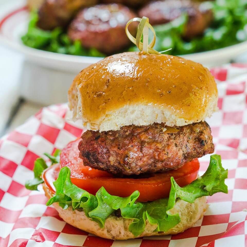 Side view of a hamburger that\'s been stuffed with Pepperjack cheese and pepperoncini sitting over a slice of tomato and lettuce between a bun. A skewer secures the bun to the burger. over a red checked cloth.
