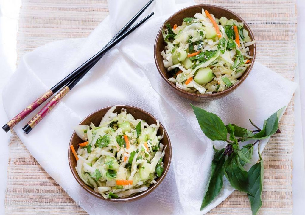 Top view of two wooden bowls heaped with Thai Basil Cabbage Slaw dotted with cucumbers sitting on a white napkin. Chopsticks and that basil sit off to the side.