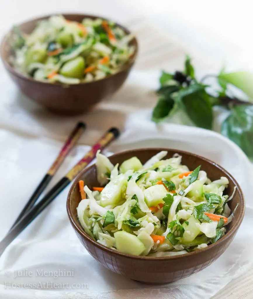 Angle view of two wooden bowls heaped with Thai Basil Cabbage Slaw dotted with cucumbers and jalapenos sitting on a white napkin. Chopsticks and that basil sit off to the side.