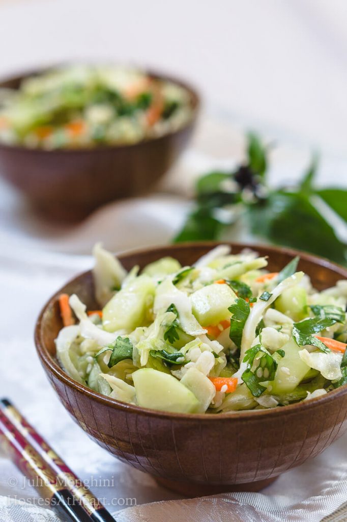 A wooden bowl heaped with Thai Basil Cabbage Slaw dotted with cucumbers and jalapenos. A second bowl sits to the back and Thai basil sits next to it.