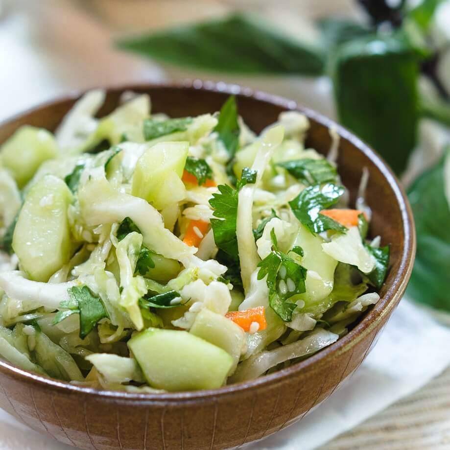 A brown bowl filled with Thai Basil Cabbage Slaw filled with cucumbers, Thai basil, carrots, cilantro, jalapenos. Thai basil sits in the background.
