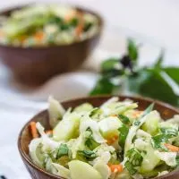 A wooden bowl heaped with Thai Basil Cabbage Slaw dotted with cucumbers and jalapenos. A second bowl sits to the back and Thai basil sits next to it.