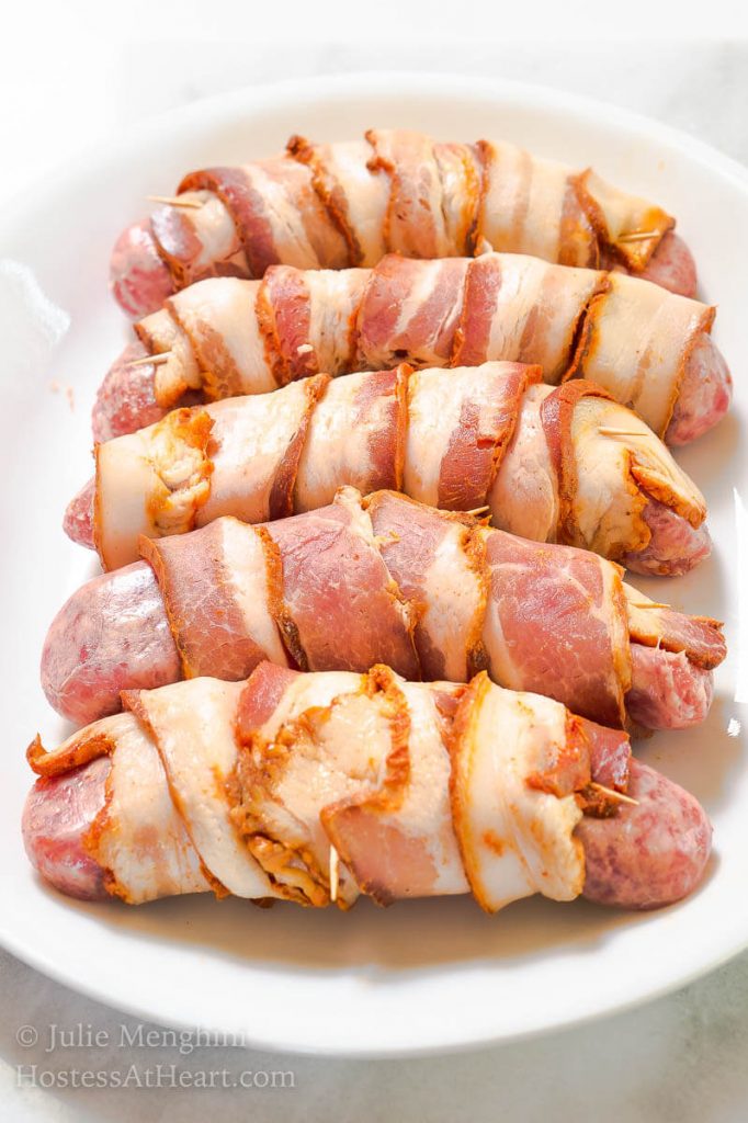 Photo of 5 brats wrapped in bacon that\'s been secured with toothpicks sitting on a white platter waiting to be cooked.