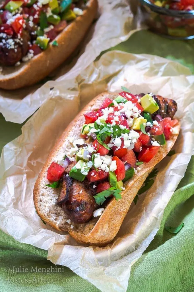 Side angle photo of a bacon-wrapped brat mounded with an avocado pico sitting in parchment paper.