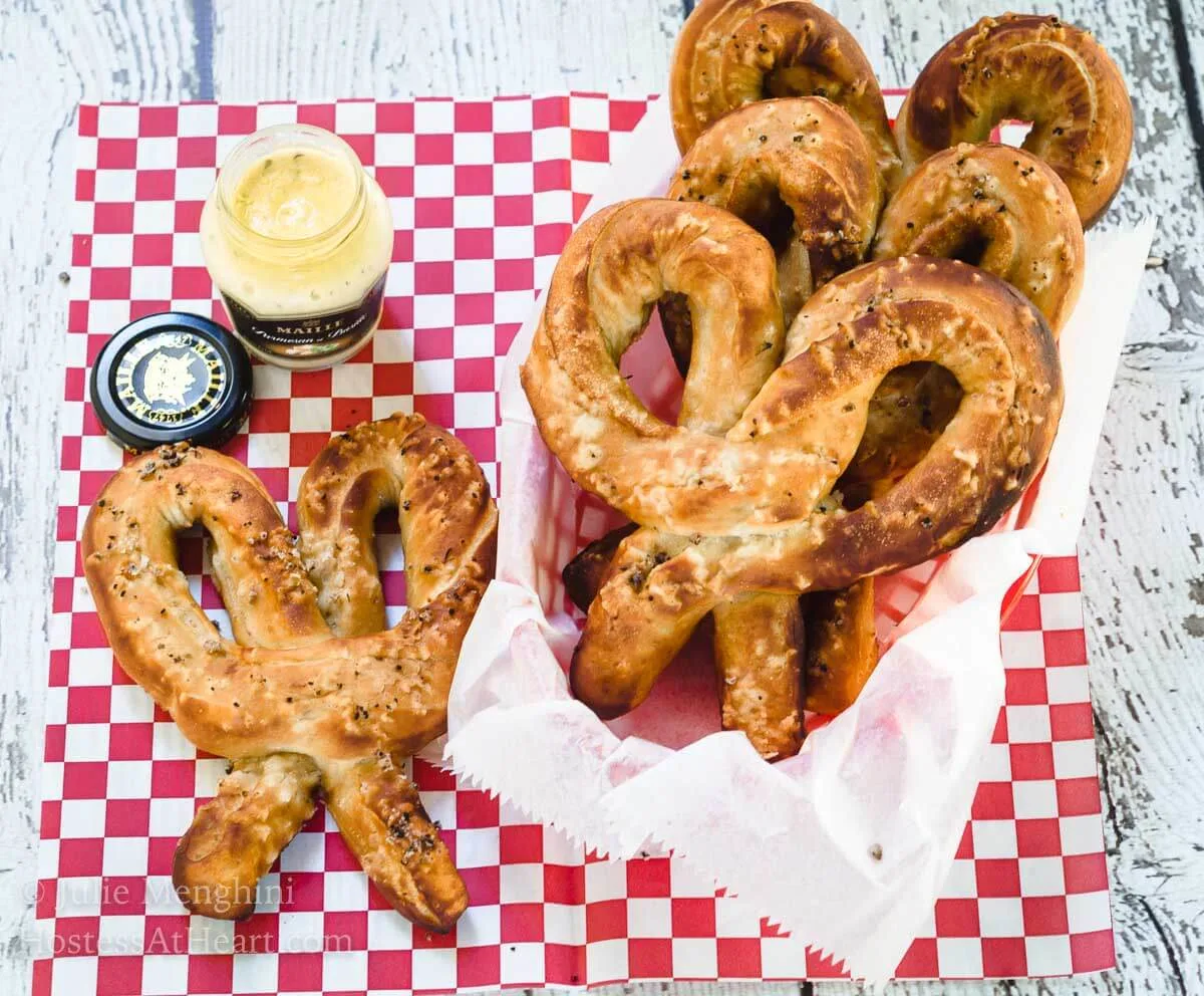 Soft pretzels and a jar of mustard sit on a red checked paper napkin. A basket of pretzels sits off to the side. 