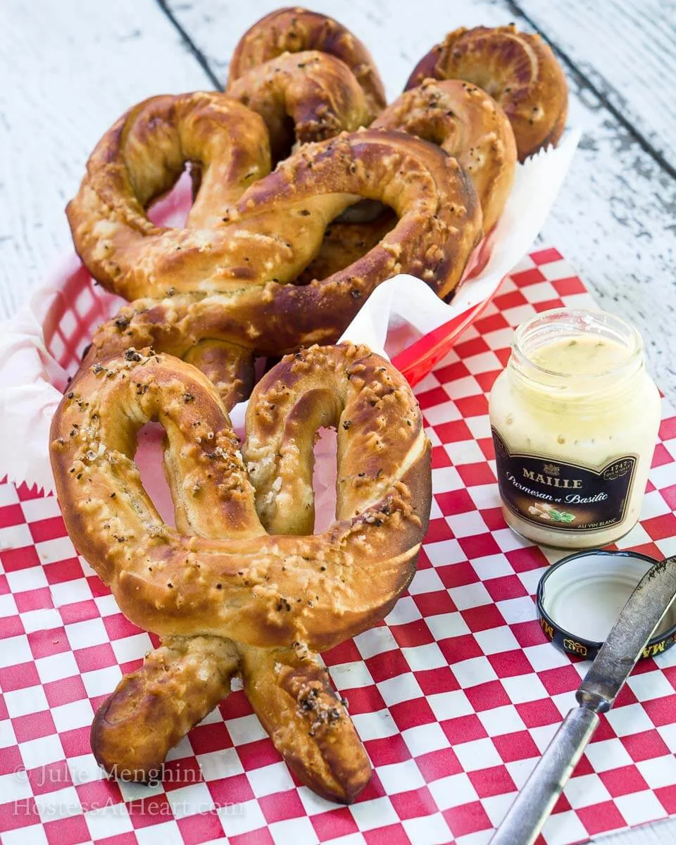 Soft pretzels and a jar of mustard sit on a red checked paper napkin. A basket of pretzels sits in the background. A spreading knife sits next to the jar.