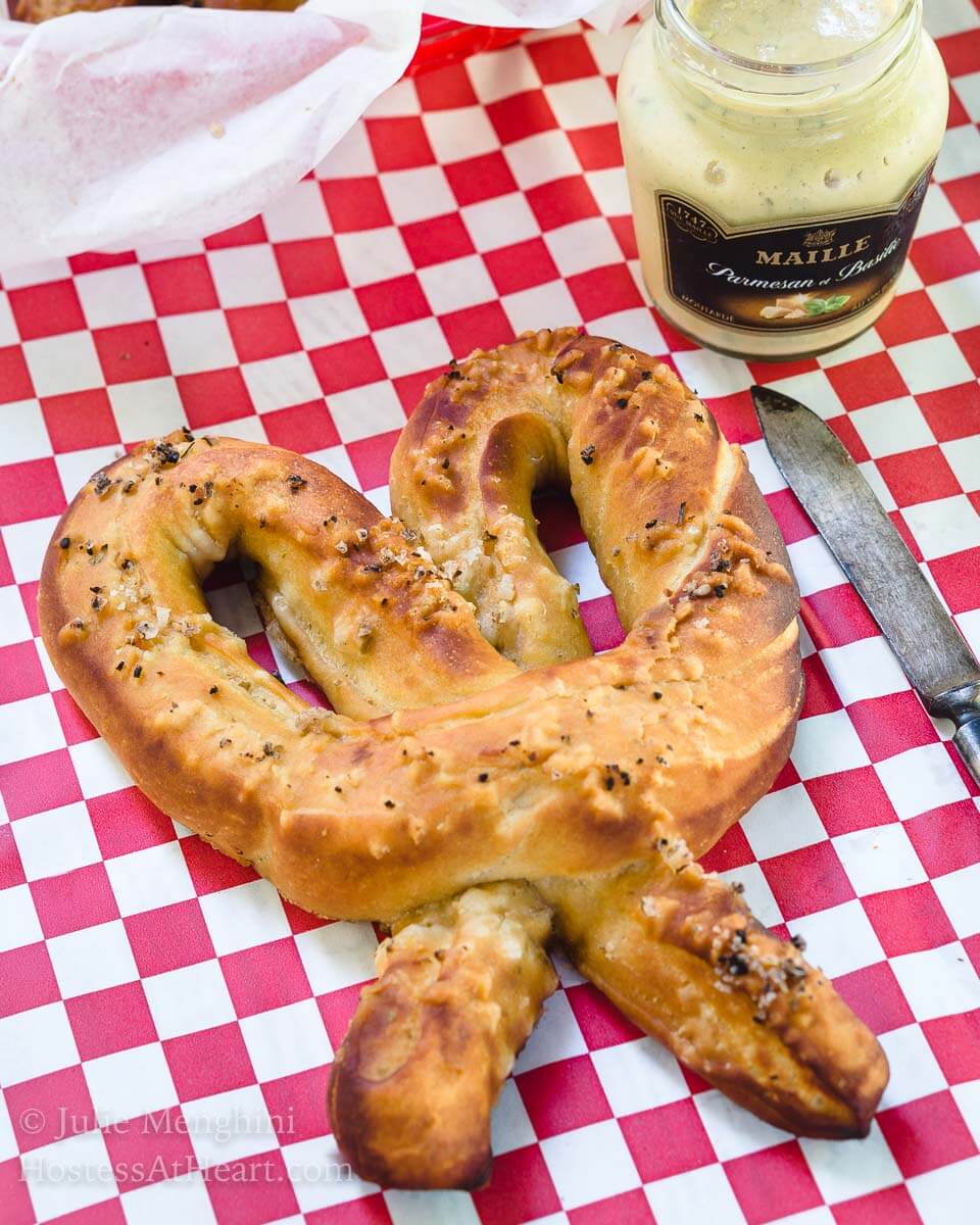 A large soft pretzel sits on a red checked paper napkin next to a spreading knife. A jar of mustard sits in the background,