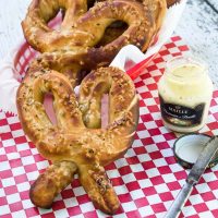 Soft pretzels and a jar of mustard sit on a red checked paper napkin. A basket of pretzels sits in the background. A spreading knife sits next to the jar.